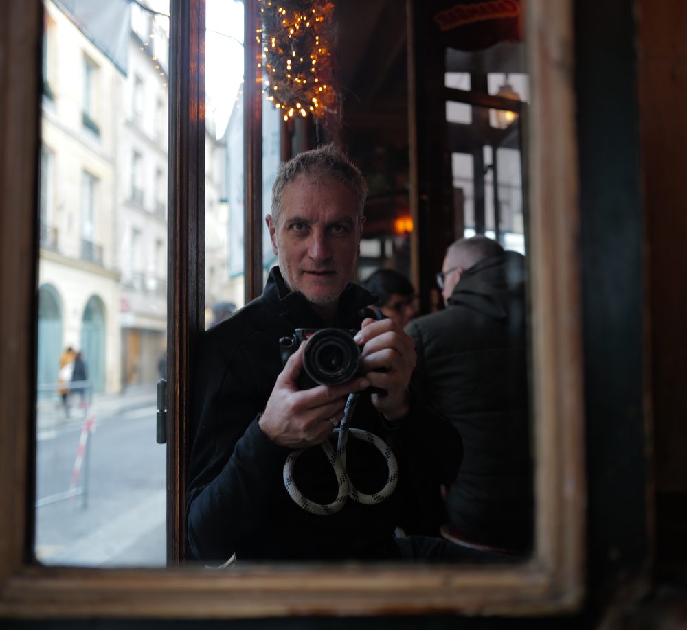 Daniel Goron reflected in a mirror hung in a Paris café window, a Leica held at his chest, the street visible through the glass beside him.